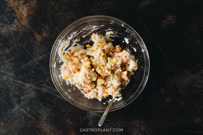 A mixing bowl with mashed potato, carrot, onion, celery, and corn