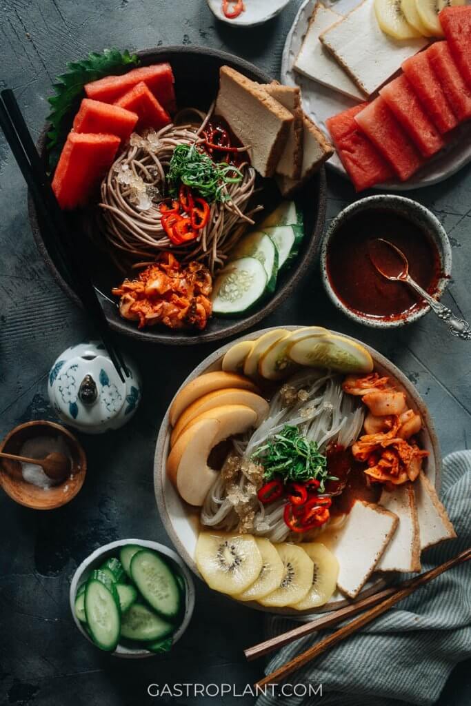 Two bowls of Korean vegan chilled noodles (mul naengmyeon) with watermelon, kimchi, kiwi, and tofu