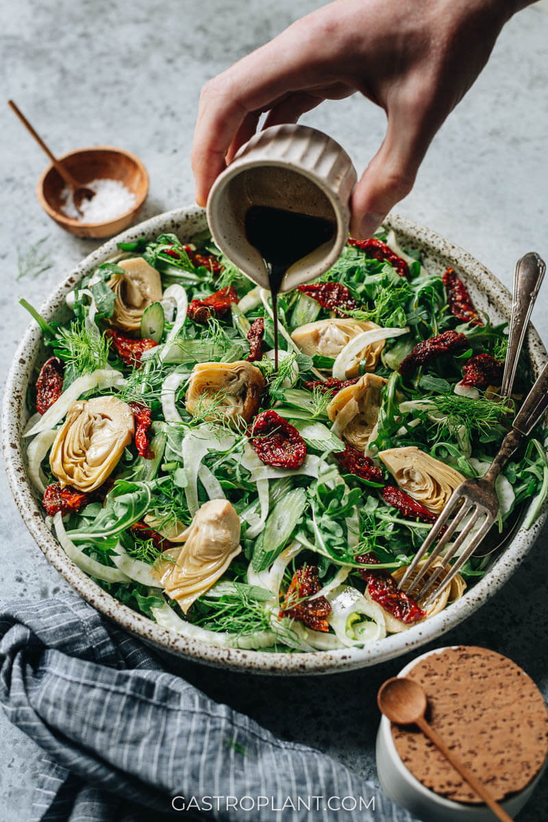 Pouring salad dressing onto easy fennel arugula salad with tomatoes and artichokes