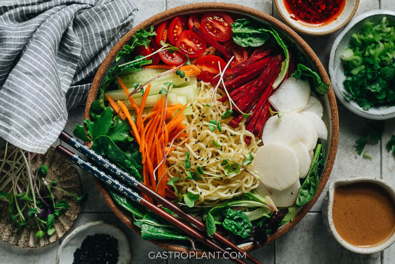 A bowl of fresh noodles with greens, beet, cucumber, carrot, radish, and a tangy nutty sesame sauce