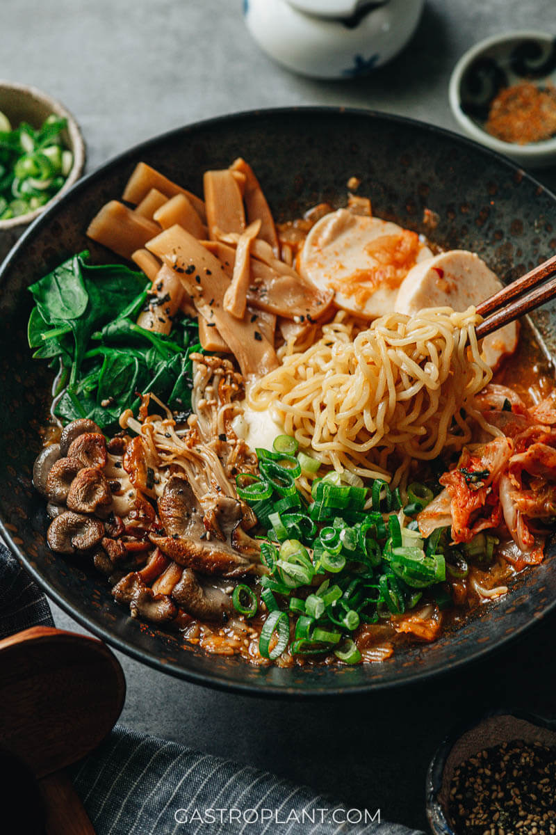 Chopsticks pulling ramen noodles from a black bowl with spicy broth