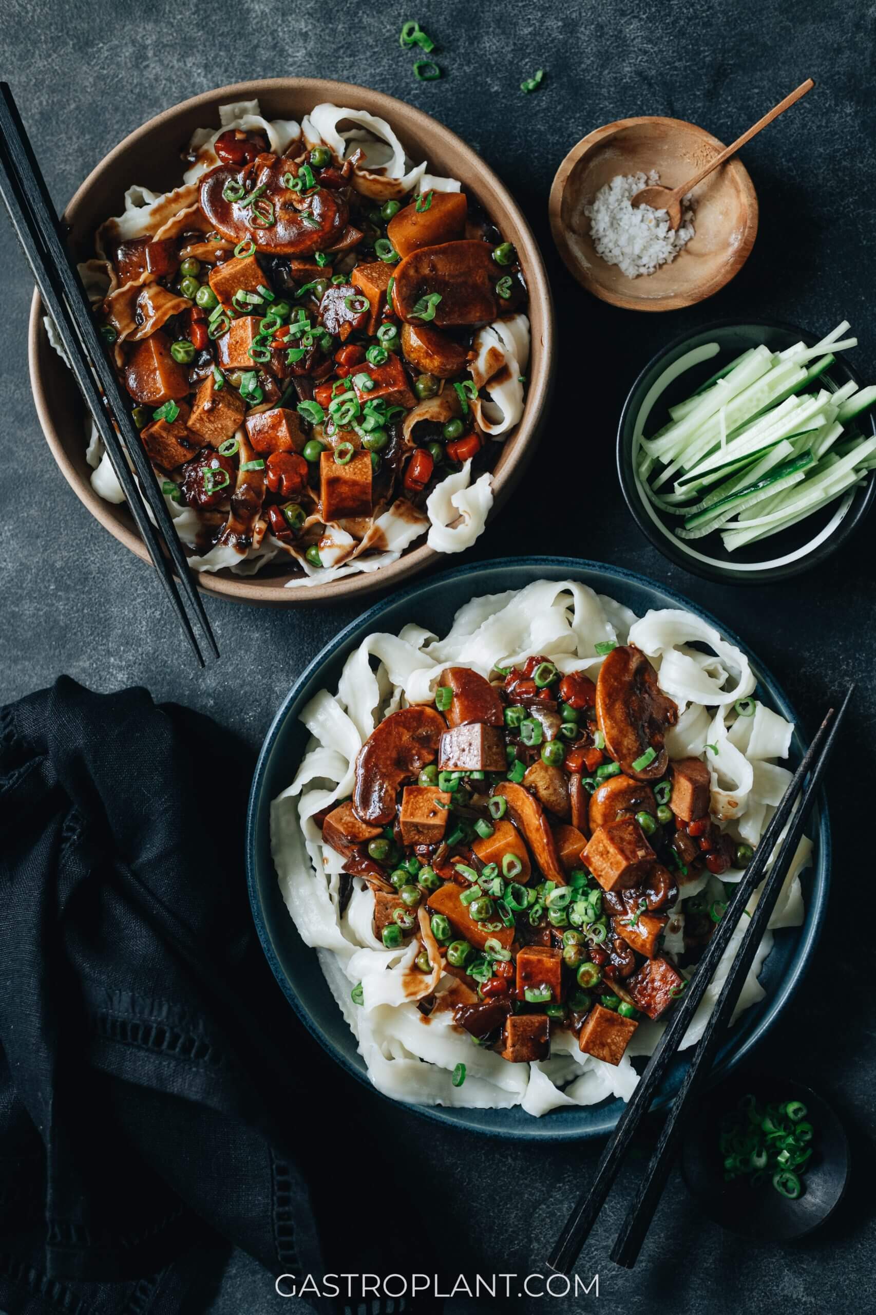 Two bowls of Korean black bean sauce noodles, fresh veggies, and sliced cucumber garnish